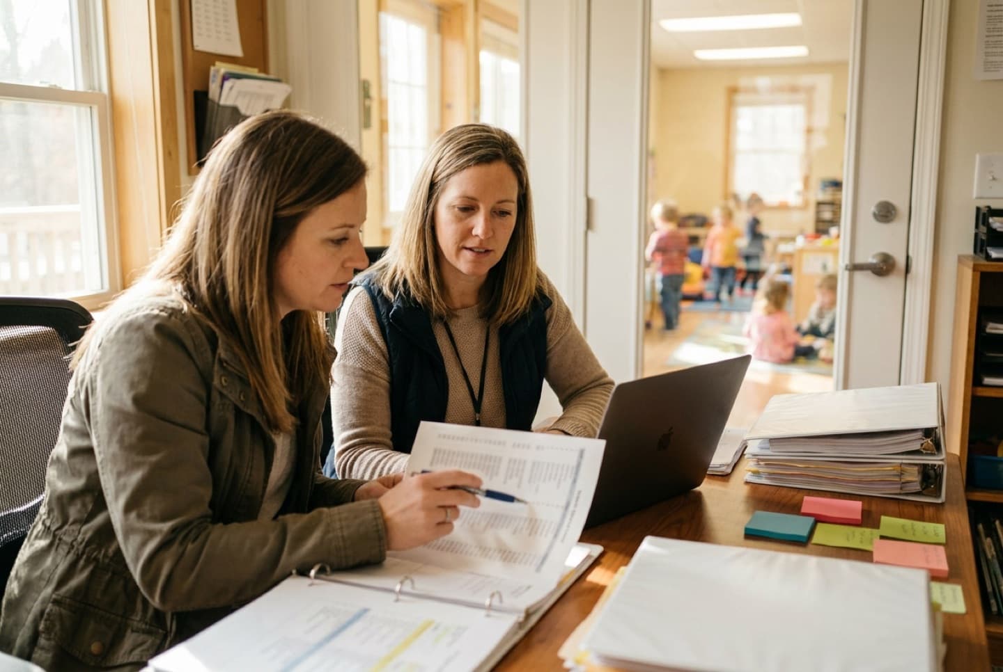 Preschool director and office administrator checking a printed family list against a laptop.